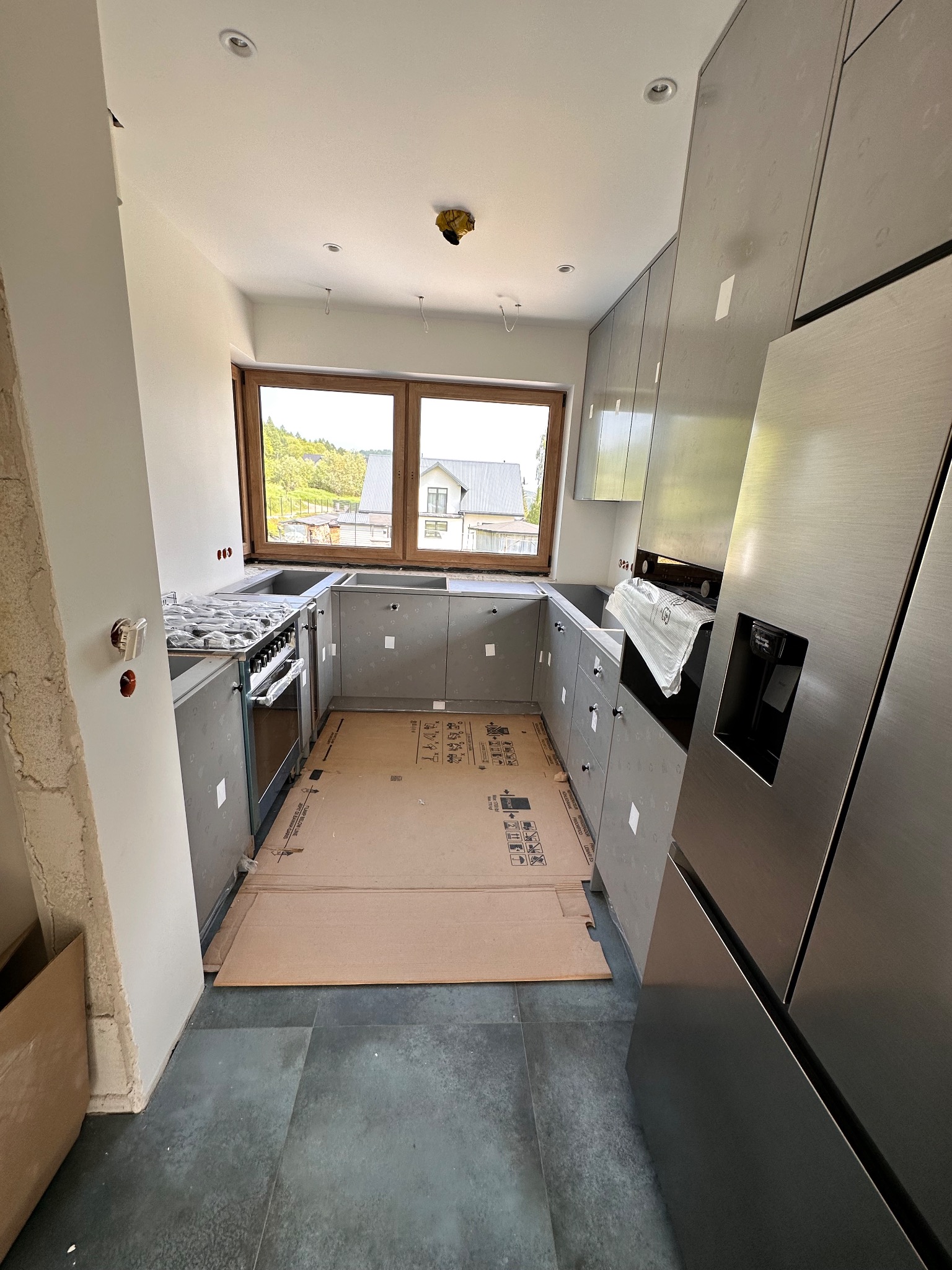 Wnętrze kuchni w trakcie montażu: szare szafki kuchenne, stalowa lodówka, okno z widokiem na zabudowę jednorodzinną, podłoga z ciemnych płytek, zabezpieczona tektura na podłodze.