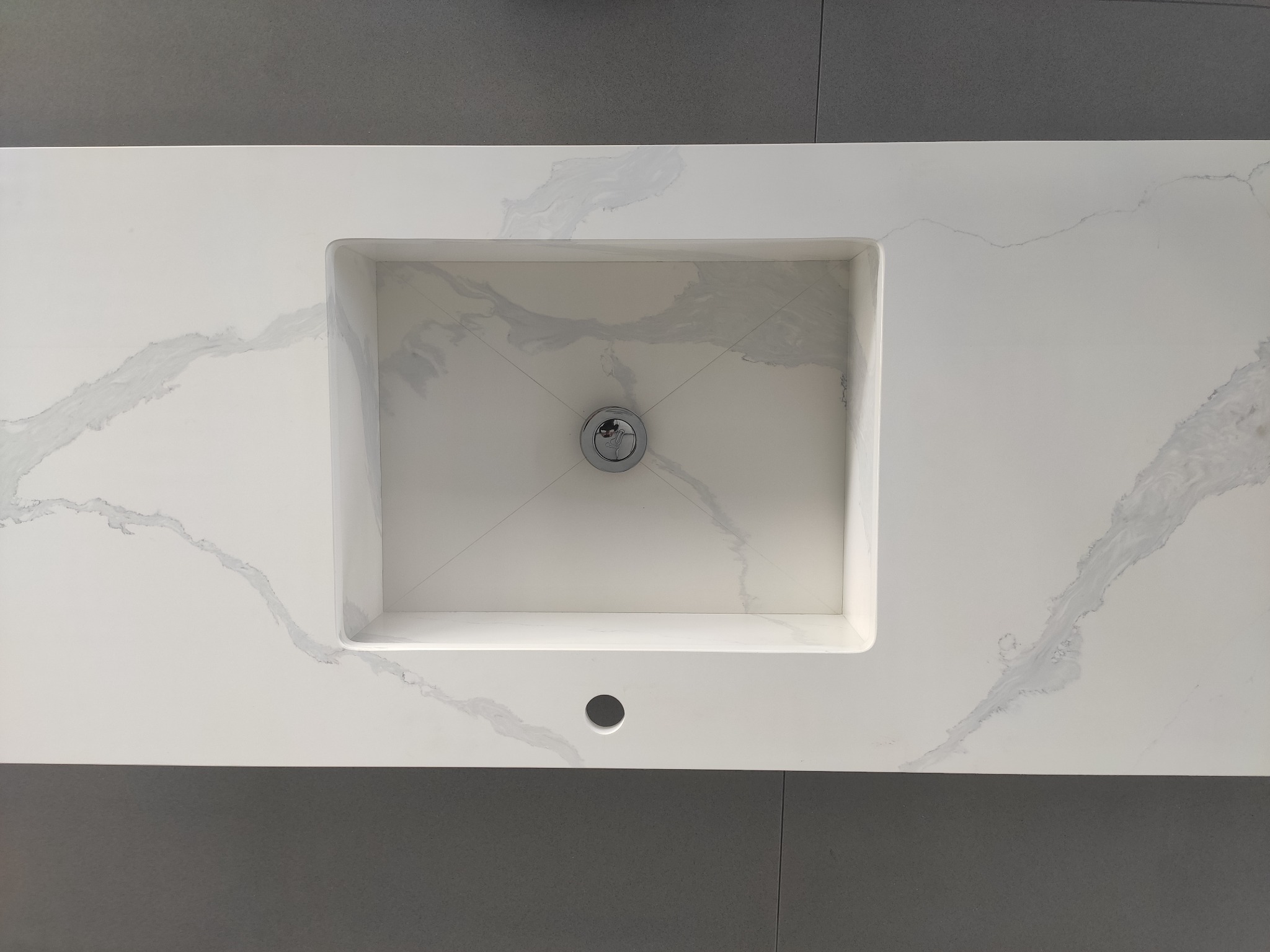 Biały blat z konglomeratu z wpuszczoną, prostokątną umywalką i chromowanym odpływem, widoczne szare żyłki wzoru na powierzchni, szara ściana w tle.