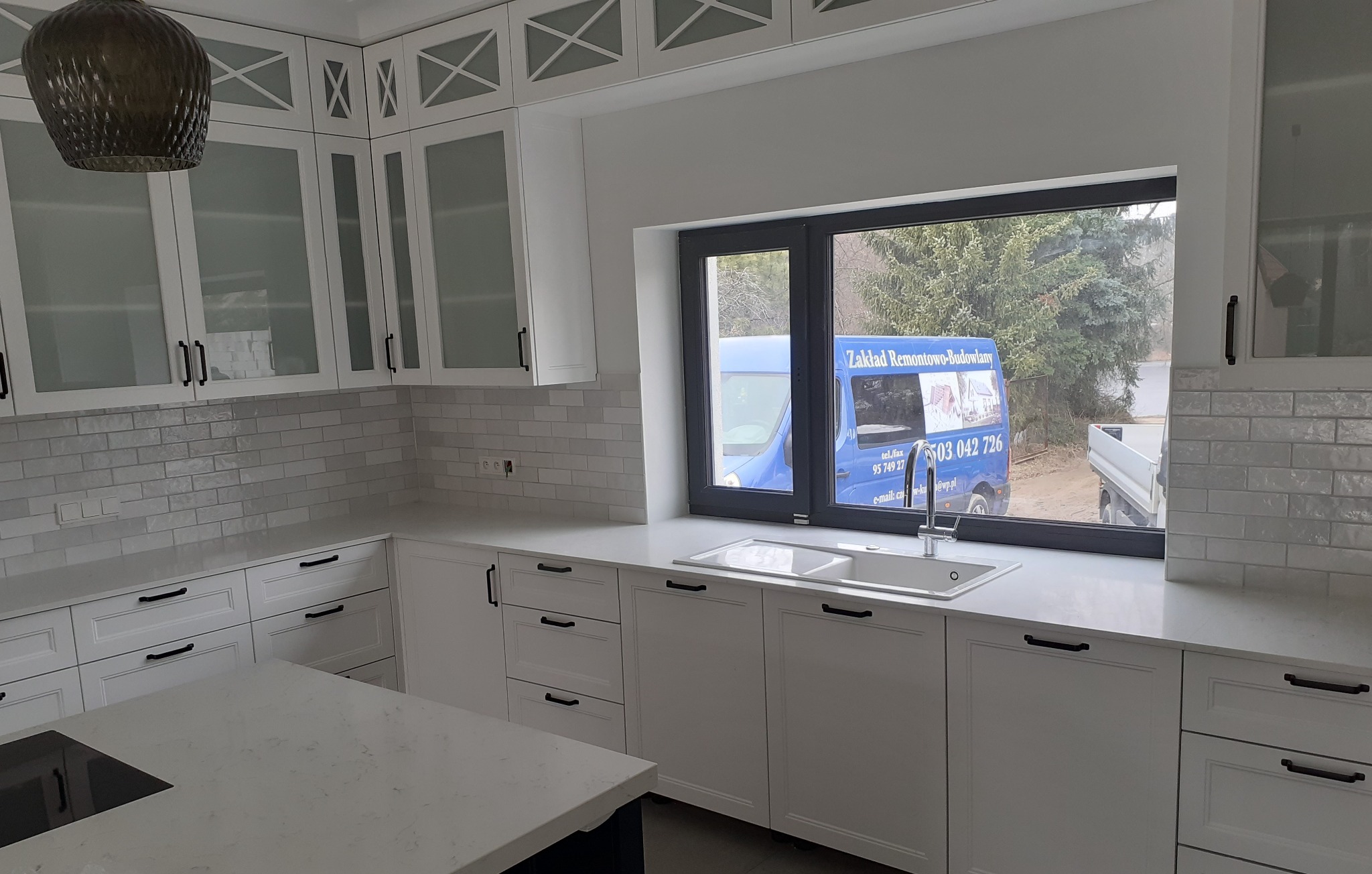 Jasna kuchnia z białymi szafkami i blatem, widok z okna na samochód firmy remontowej.