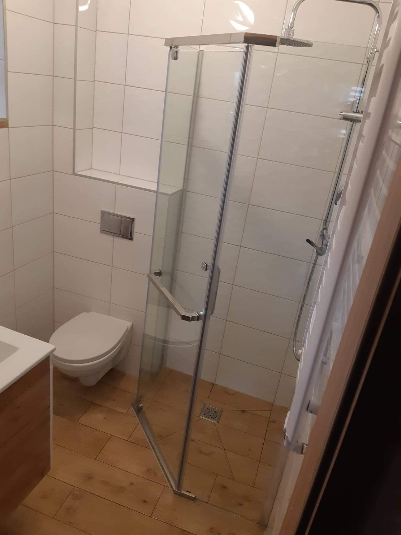 Nowoczesna kabina prysznicowa ze szklanymi drzwiami, zamontowana w narożniku łazienki wyłożonej białymi płytkami i drewnopodobną podłogą, widoczny odpływ liniowy i wisząca toaleta.