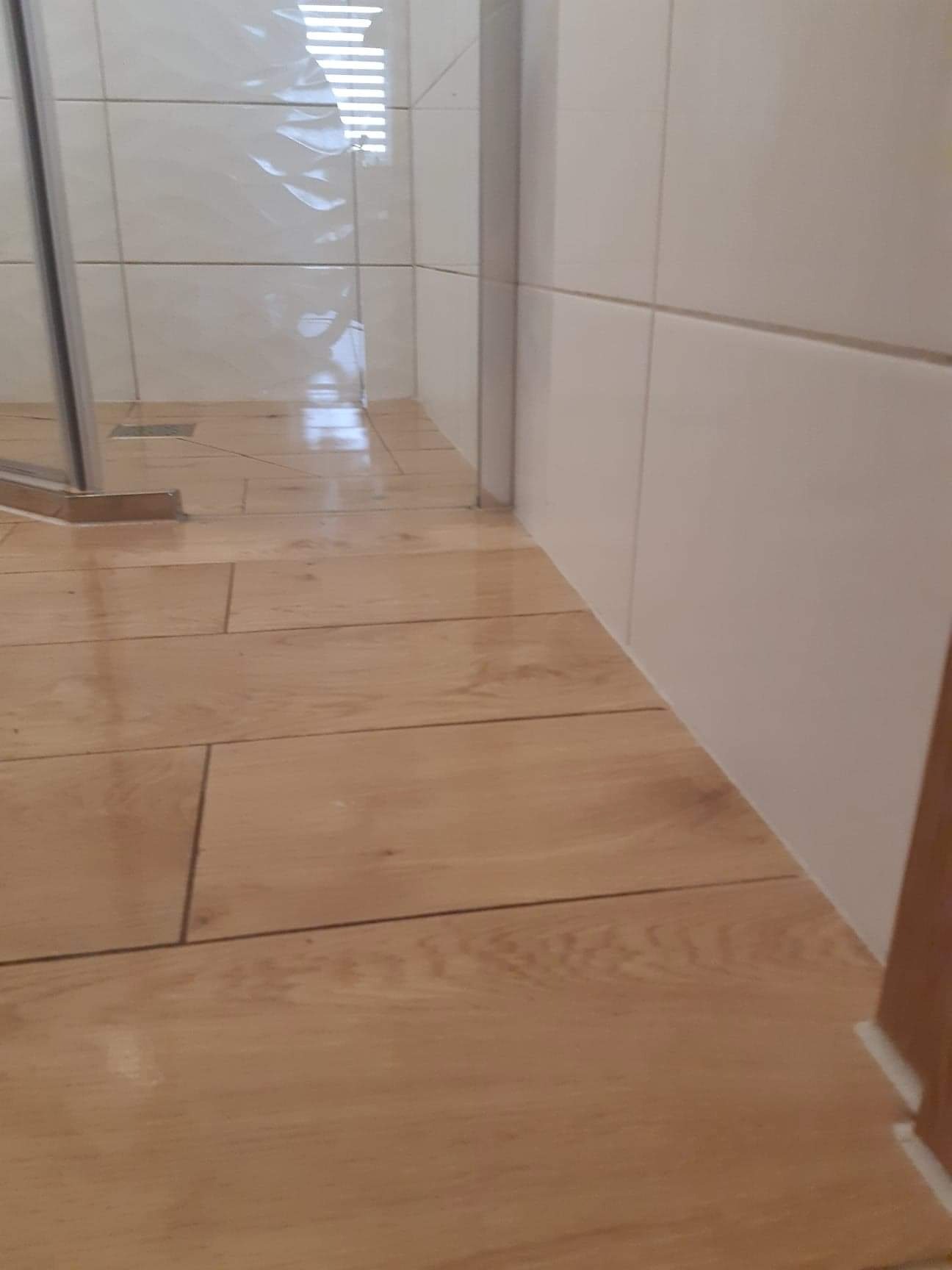 Jasne, drewnopodobne płytki podłogowe w kabinie prysznicowej z białymi płytkami ściennymi, widoczne odpływ liniowy i fragment szklanej ścianki kabiny.