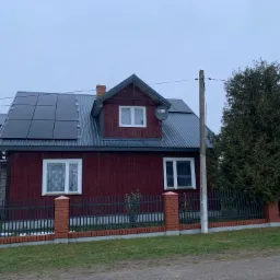 Ciemnoczerwony dom jednorodzinny z szarym dachem pokrytym panelami słonecznymi, widoczne ogrodzenie z ceglanymi słupkami i drzewo iglaste z prawej strony.