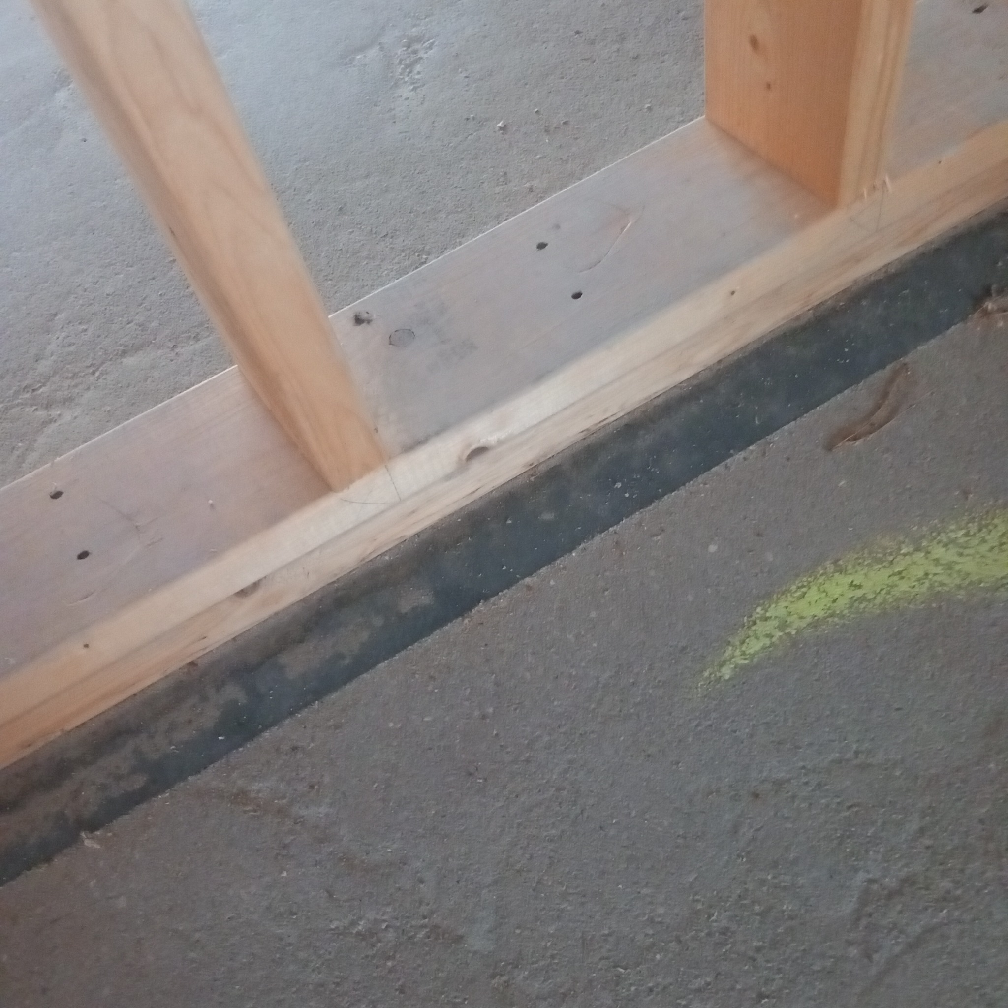 Fragment konstrukcji szkieletowej domu z widocznymi drewnianymi belkami i betonową podłogą z zielonymi śladami farby.