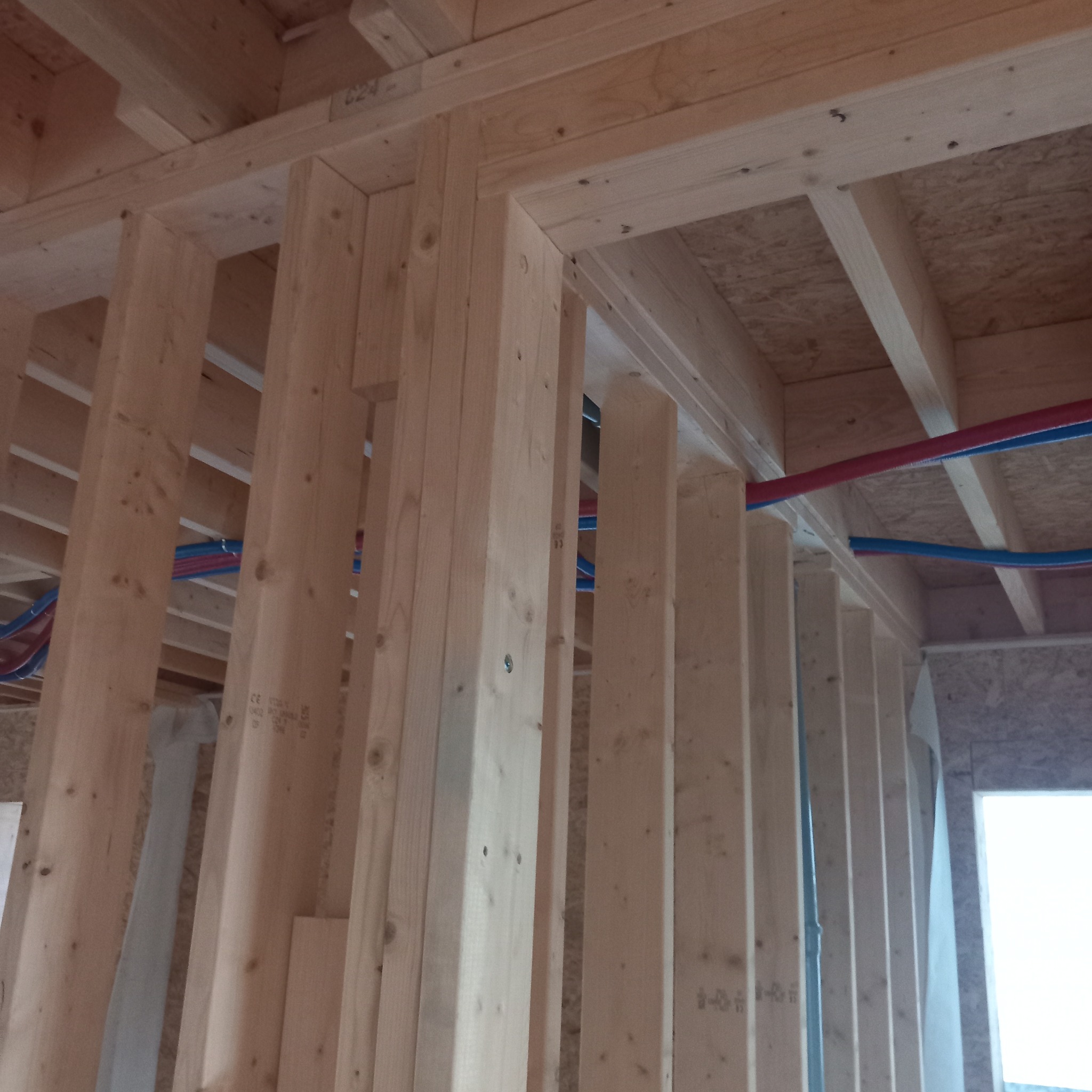 Wnętrze domu w trakcie budowy, widoczne drewniane elementy konstrukcyjne ścian i stropu, z instalacją wodną w kolorze niebieskim i czerwonym, oraz fragment okna.