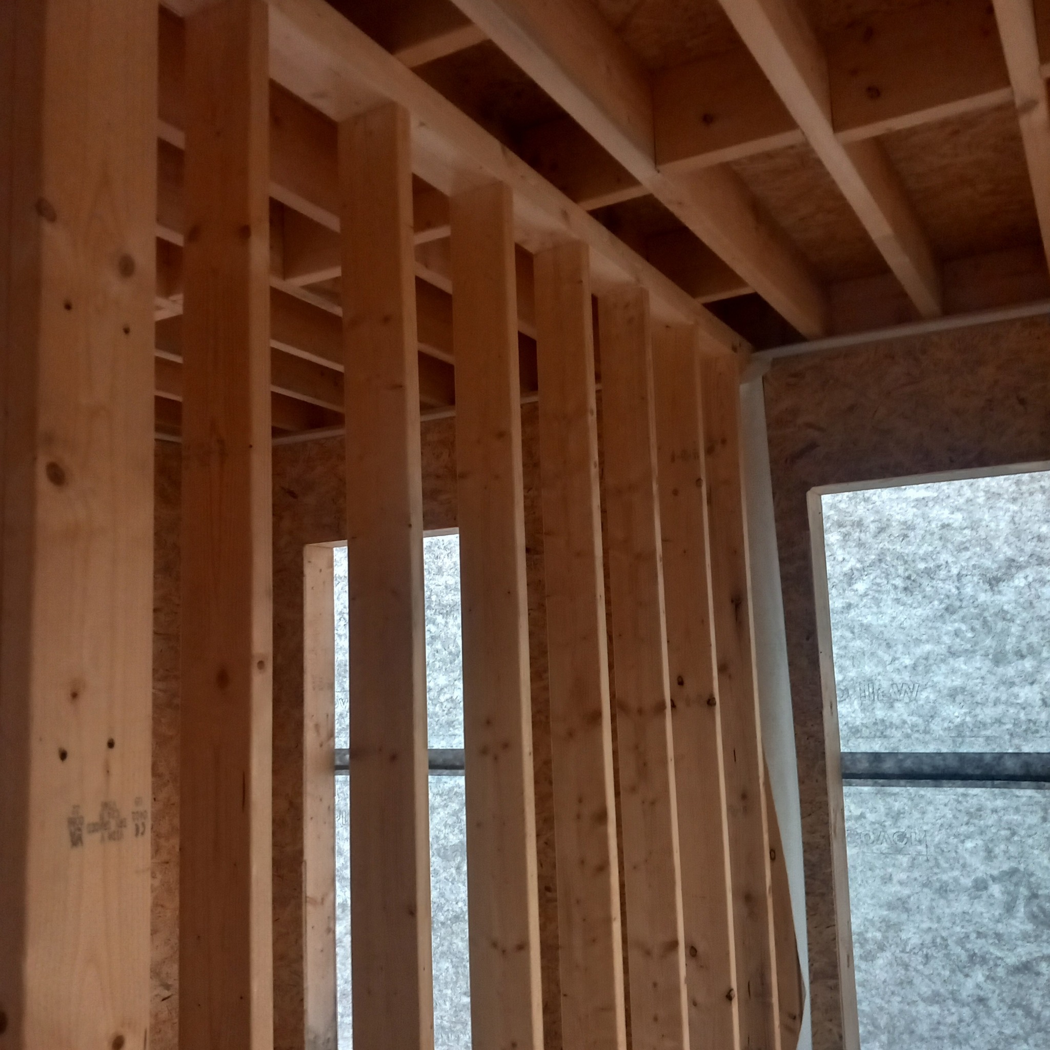 Wnętrze w trakcie budowy domu szkieletowego, widoczne drewniane belki konstrukcyjne ścian i stropu, płyty OSB oraz membrana wiatroizolacyjna na ścianie zewnętrznej.