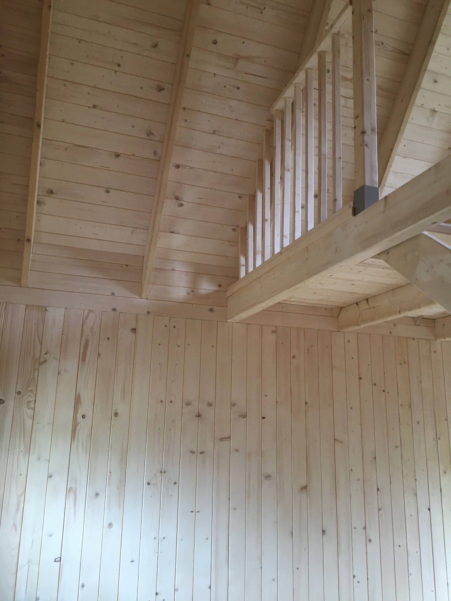 Wnętrze domu z surowego drewna, widok na antresolę z drewnianą balustradą i skośny sufit z drewnianych desek.