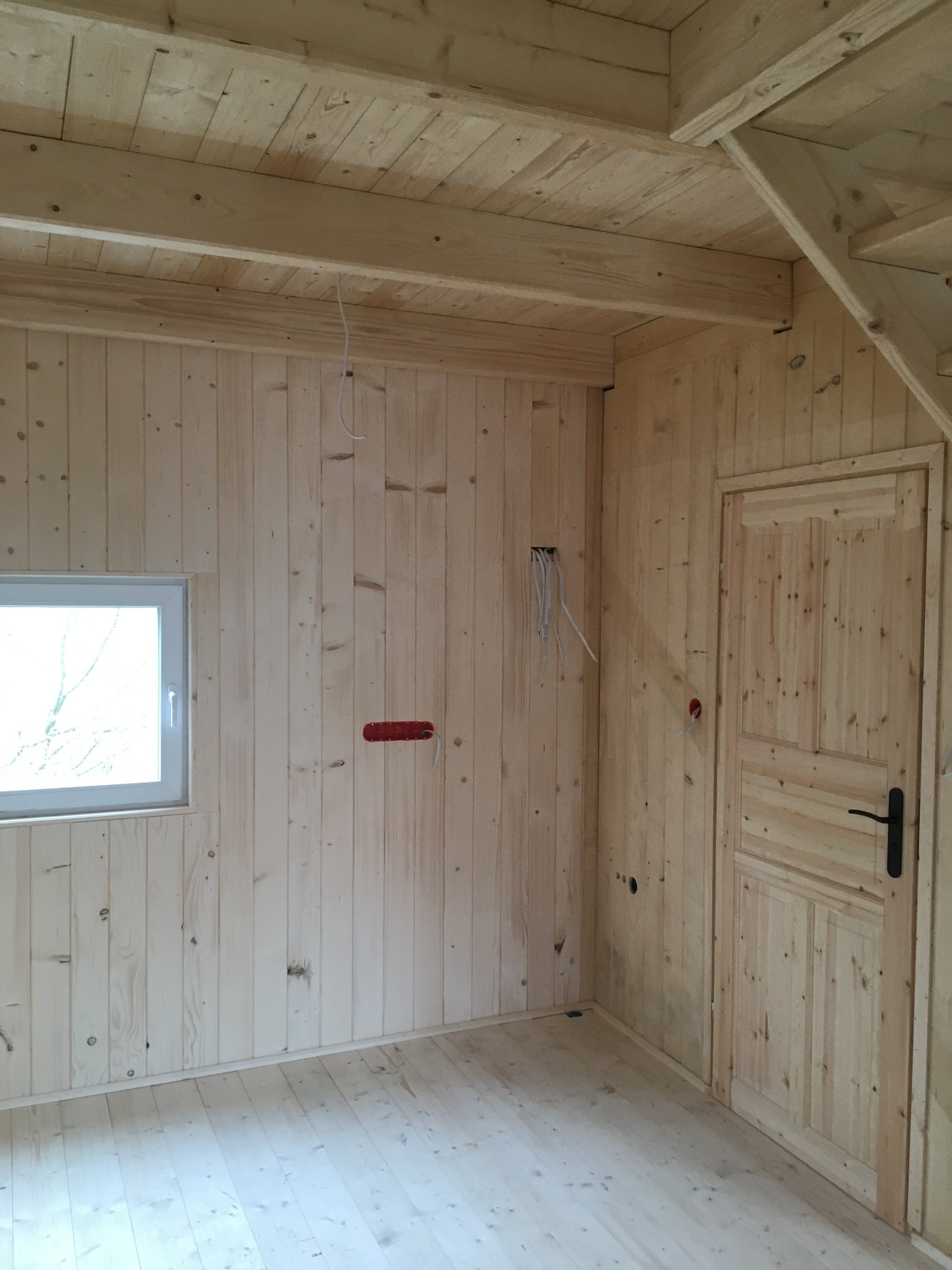 Wnętrze domu w konstrukcji szkieletowej, wykończone jasnym drewnem, widoczne instalacje elektryczne w trakcie montażu, okno z widokiem na zewnątrz, drewniane drzwi z czarną klamką.