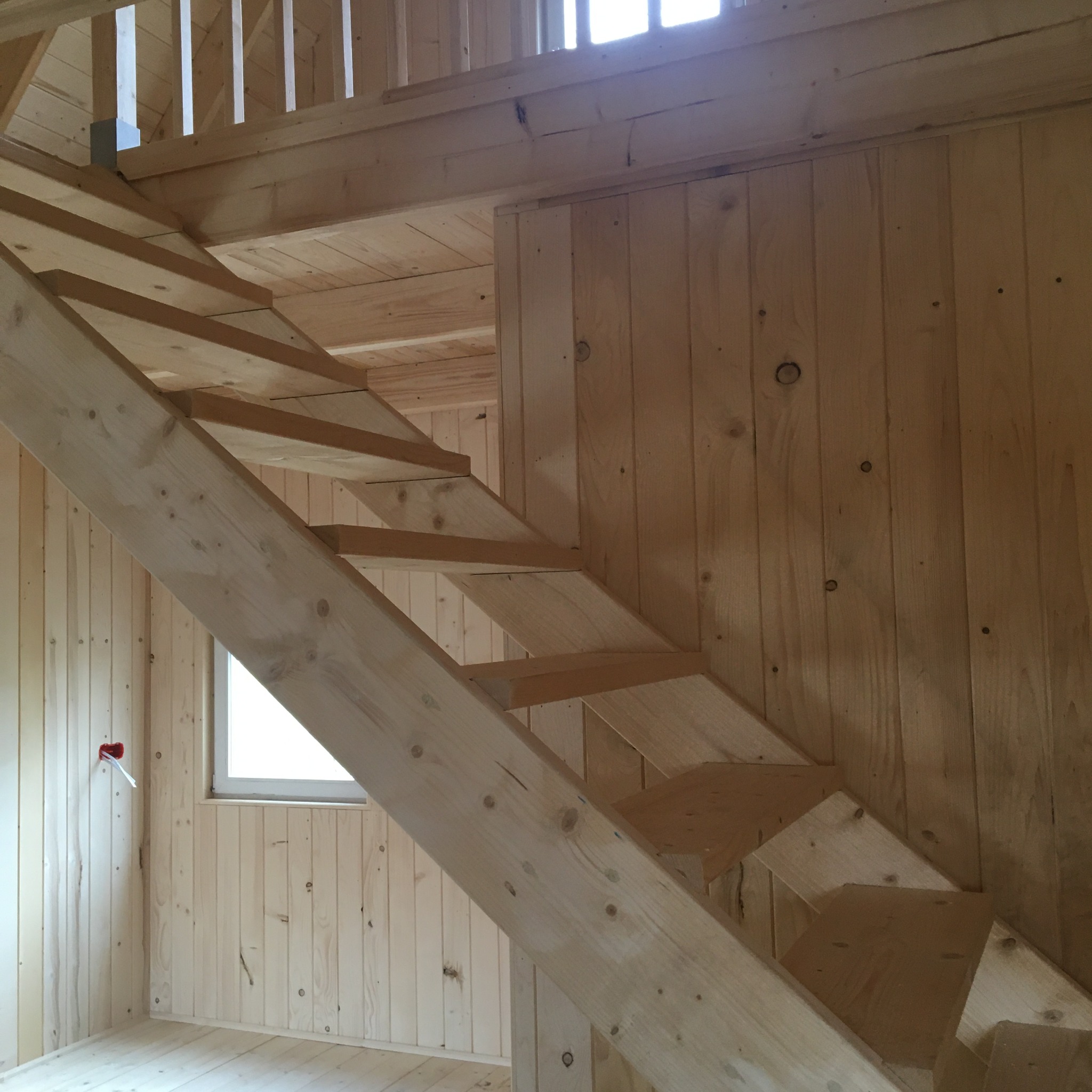 Surowe drewniane schody wewnątrz domu w konstrukcji szkieletowej, widoczne deski ścian i podłogi, małe okno w tle.