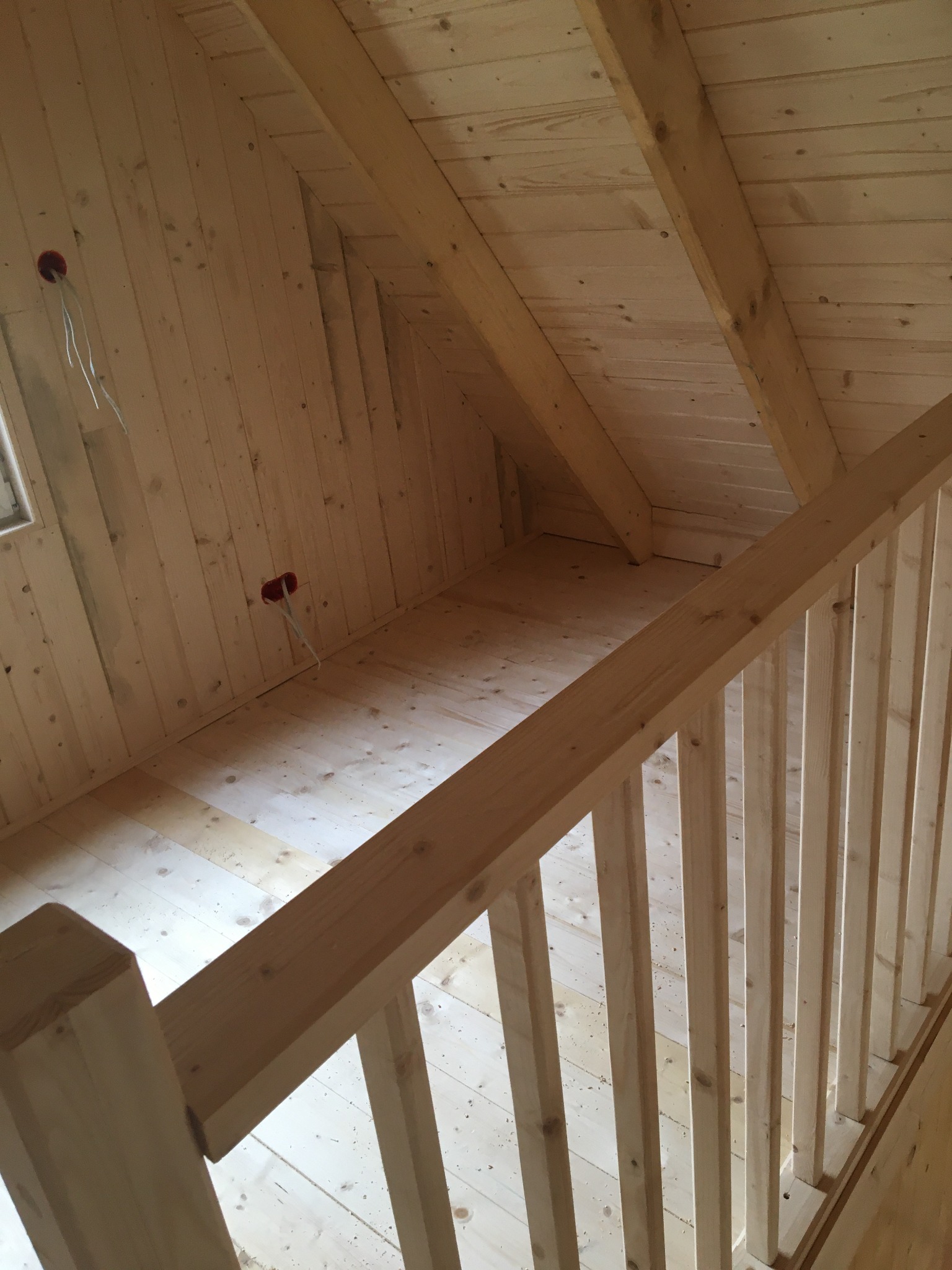 Surowe wnętrze poddasza z drewnianą konstrukcją szkieletową i schodami, widoczne gniazdka elektryczne z wystającymi kablami, surowe wykończenie.