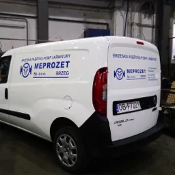 Biały samochód dostawczy z niebieskim logo i nazwą firmy 'MEPROZET' z Brzegu, zaparkowany w hali produkcyjnej.