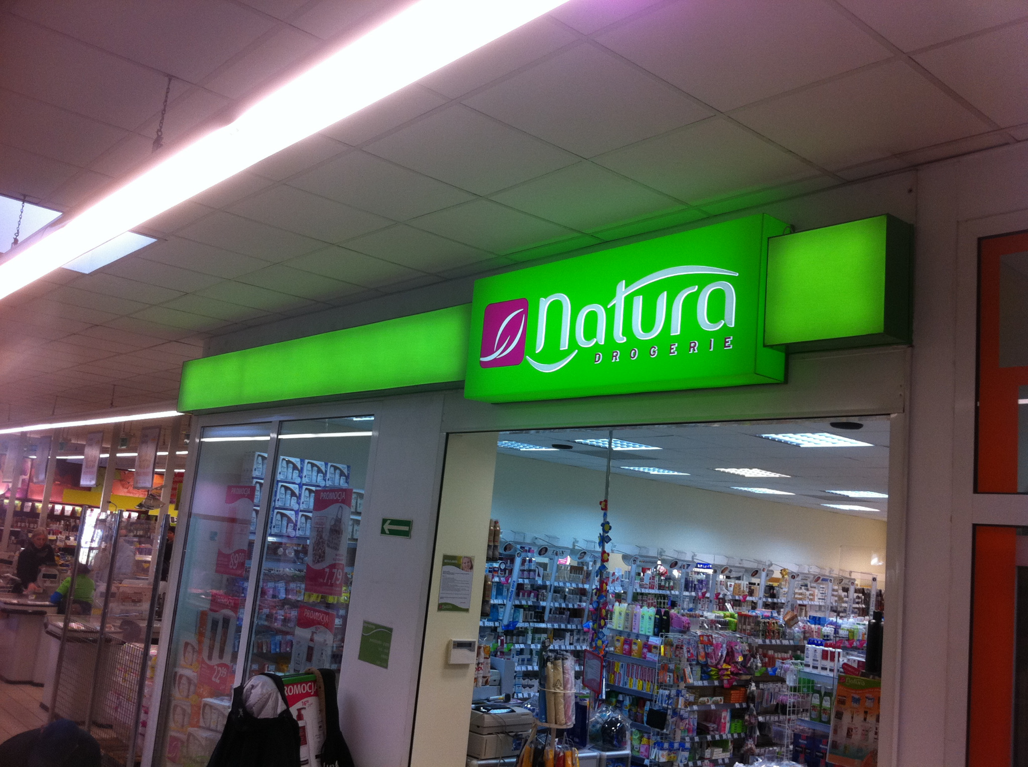 Podświetlany, zielony szyld z logo i napisem 'Natura Drogerie' nad wejściem do sklepu, widoczne półki z produktami wewnątrz.