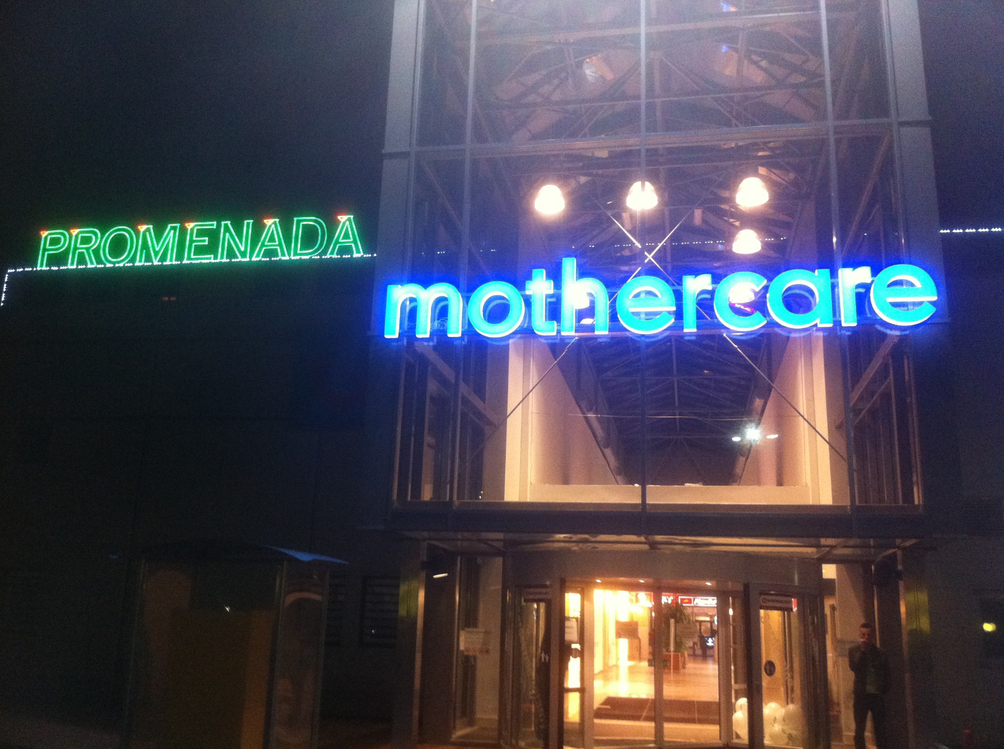 Fasada budynku z neonowym szyldem 'mothercare' podświetlonym na niebiesko i zielonym napisem 'PROMENADA' obok, widoczna część wejścia z obrotowymi drzwiami.