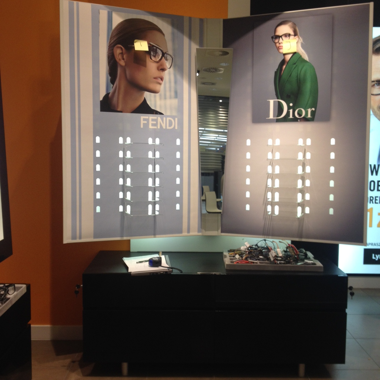 Ekspozycja okularów Fendi i Dior na podświetlanych panelach z pustymi półkami, na czarnym blacie leżą notatki i plątanina kabli.