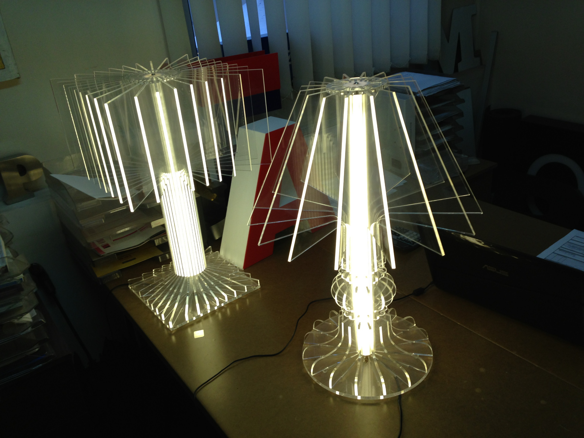 Dwie designerskie lampy stołowe z przezroczystego akrylu, podświetlane od wewnątrz, na biurku z laptopem w tle.