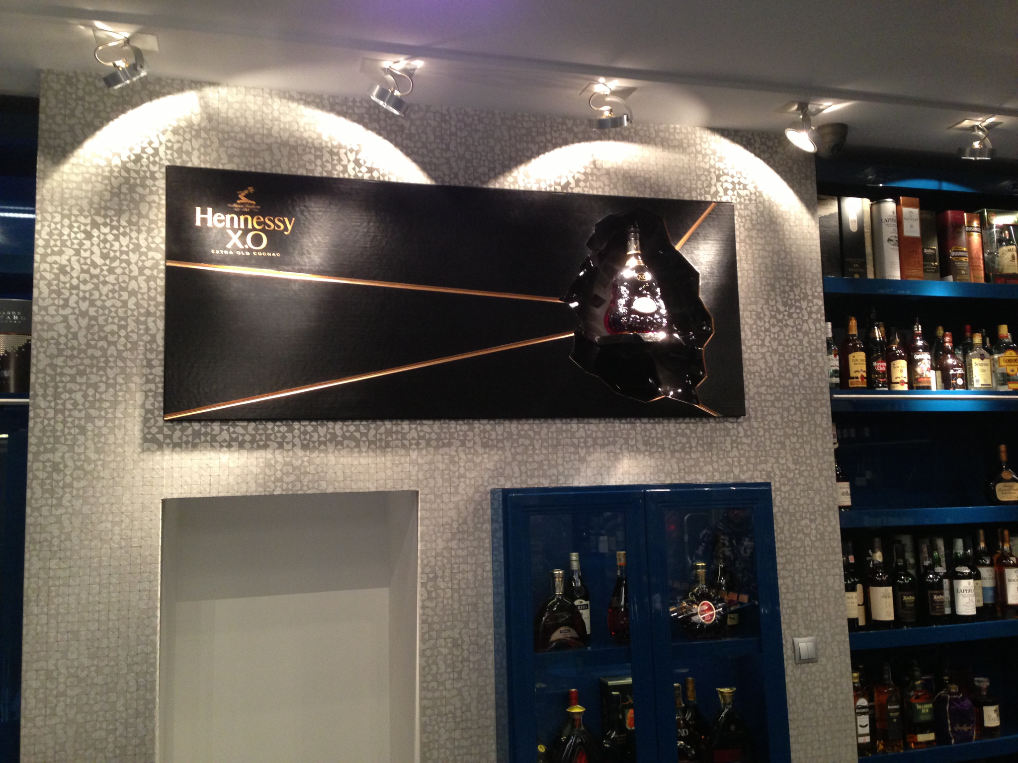 Czarny panel z podświetlonym logo Hennessy XO i butelką koniaku w nieregularnym wycięciu, umieszczony na ścianie z wzorzystą tapetą, oświetlony reflektorami punktowymi.