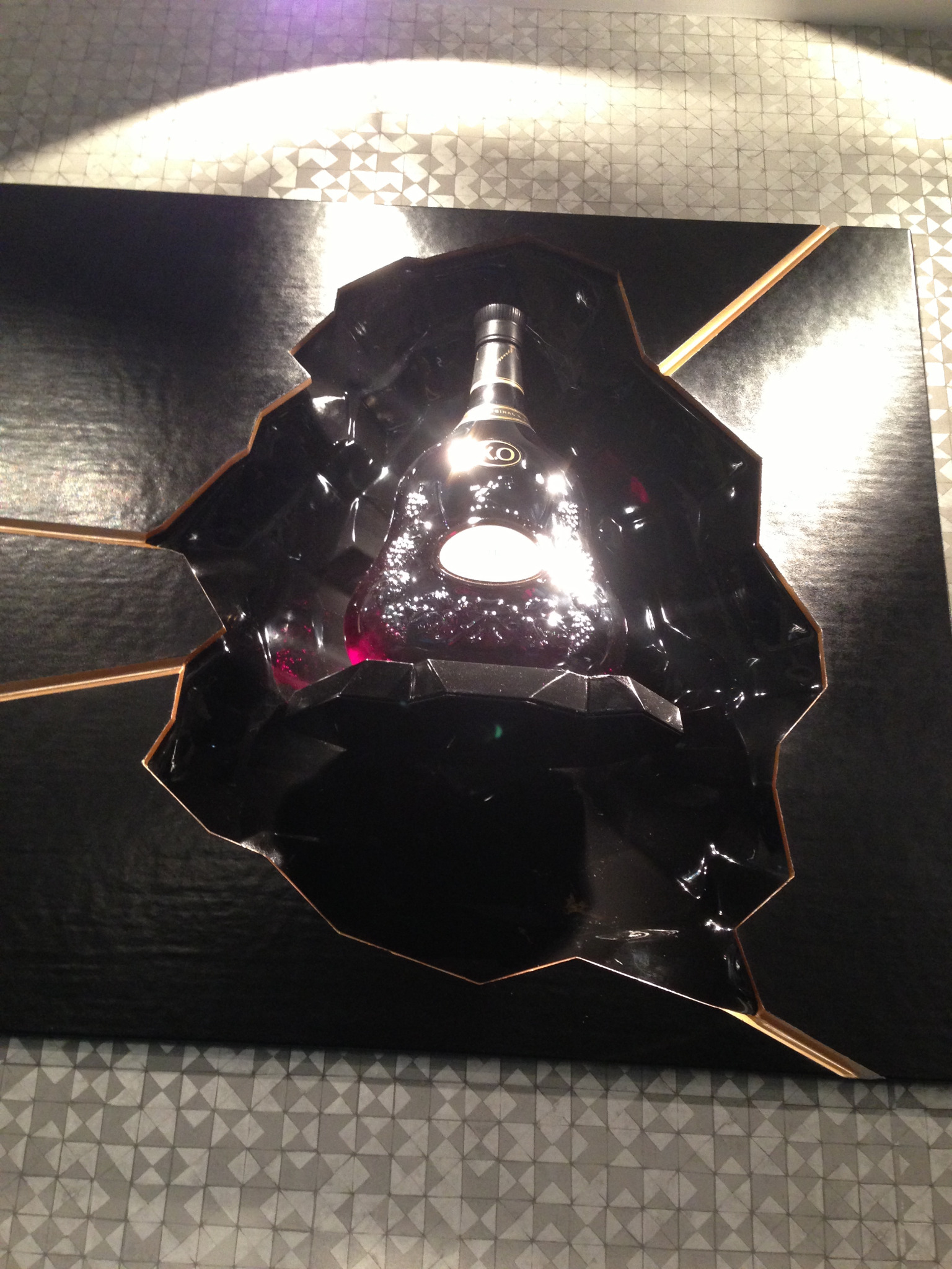 Butelka alkoholu w czarnym opakowaniu z geometrycznymi, złotymi akcentami, oświetlona punktowym światłem.