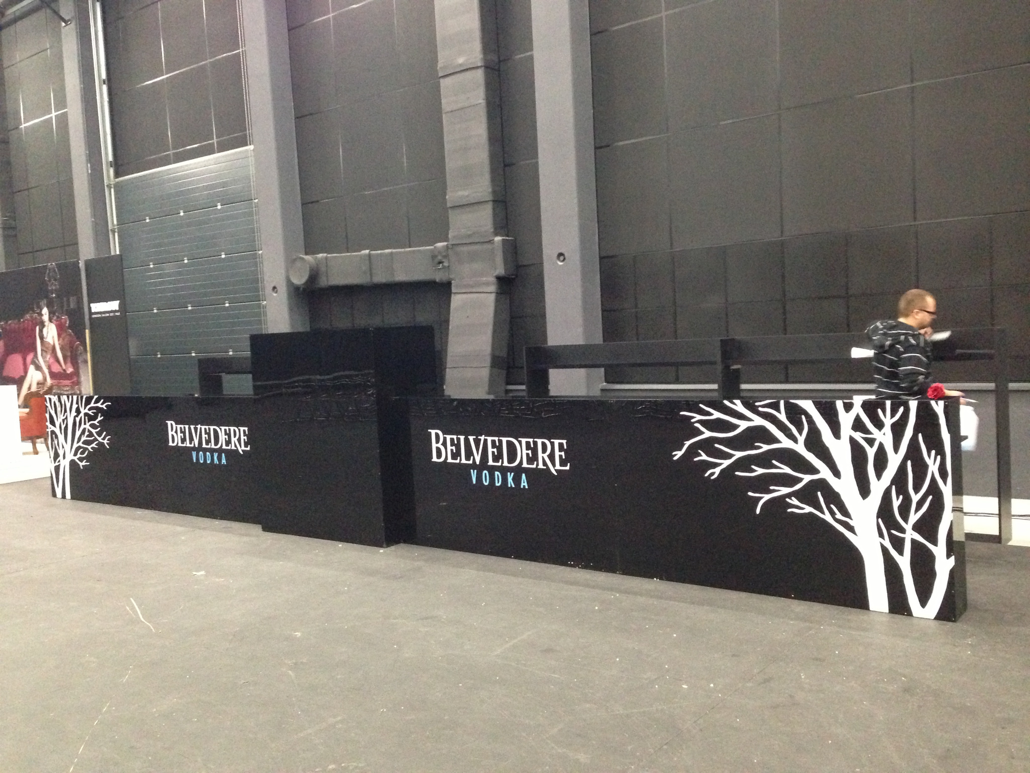 Czarny bar z białym logo i grafiką drzewa Belvedere Vodka na tle szarej ściany hali, mężczyzna w okularach stoi za barem.
