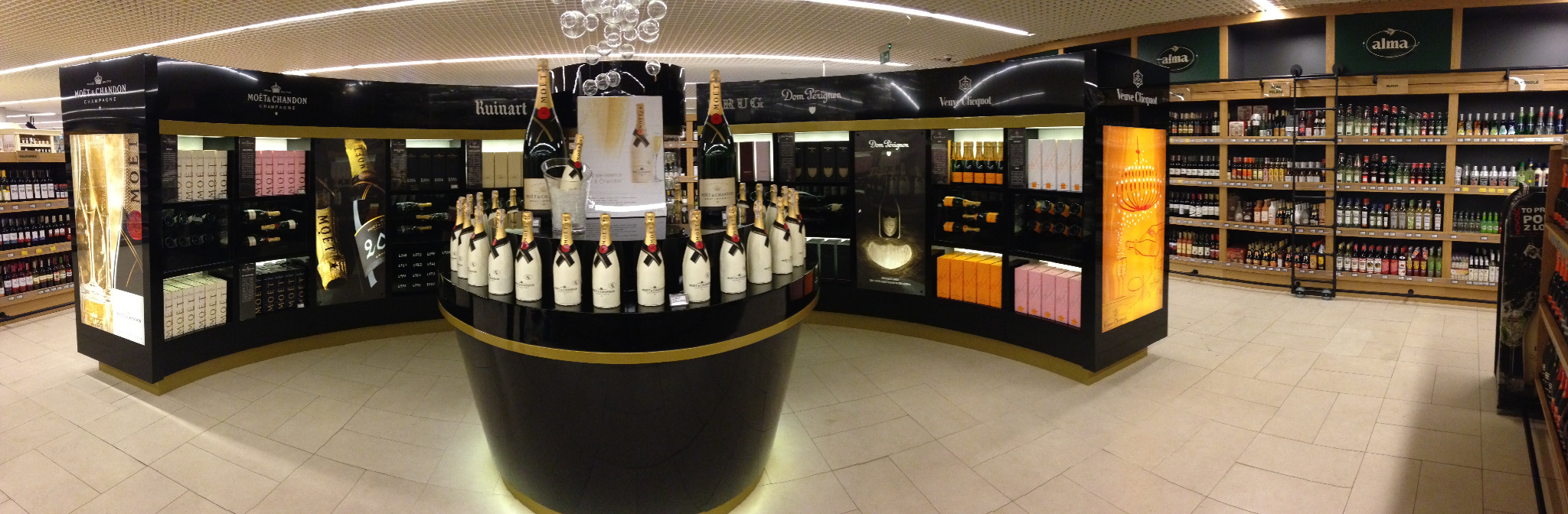 Panoramiczny widok na stoisko z szampanami w sklepie, prezentujące różne marki i modele w eleganckich, podświetlanych ekspozytorach.