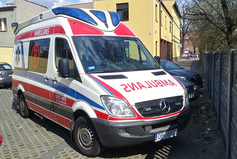 Oklejony ambulans Mercedes Sprinter z czerwono-biało-niebieskimi pasami i napisem AMBULANS, zaparkowany na brukowanym parkingu przed budynkiem. Widoczne logo firmy KOST MED i numer telefonu na boku...
