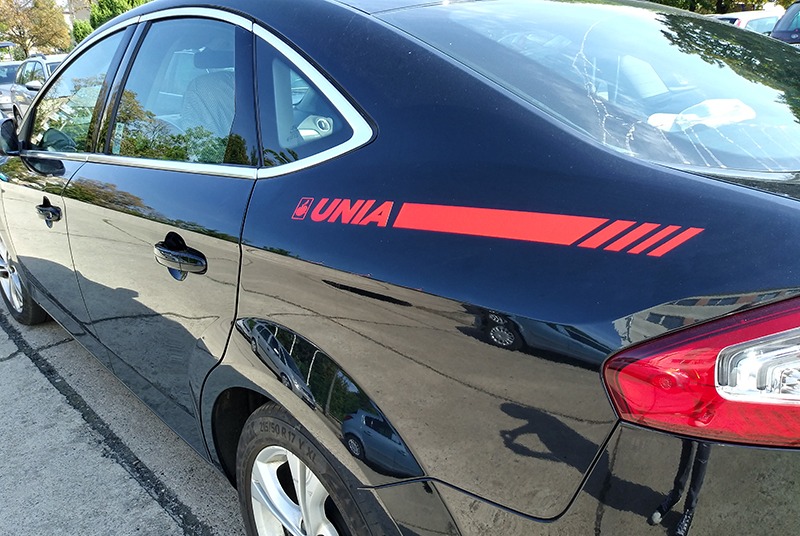 Czarny samochód osobowy z czerwonym logo i paskami 'UNIA' na tylnym błotniku, widoczne opony i fragment tylnej lampy.