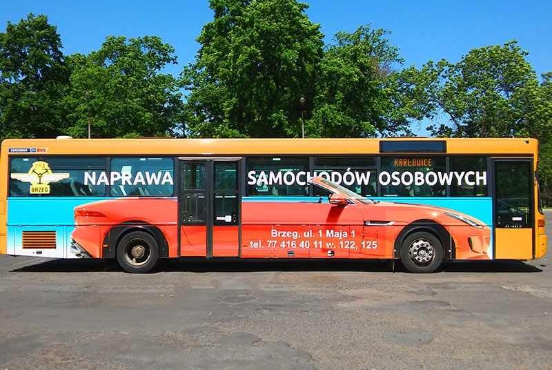 Autobus miejski w żółto-pomarańczowych barwach z reklamą serwisu samochodowego, na boku grafika przedstawiająca fragment sportowego samochodu i napis 'Naprawa Samochodów Osobowych'.