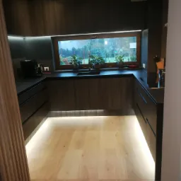 Nowoczesna kuchnia z ciemnego drewna z podświetleniem LED pod szafkami i blatem, okno z roślinami doniczkowymi, drewniana podłoga