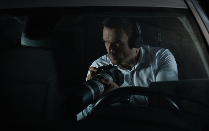 Mężczyzna w białej koszuli i słuchawkach, siedzący w samochodzie w Gdyni, trzyma aparat z dużym obiektywem, prawdopodobnie robiąc zdjęcia.