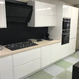 Biała, nowoczesna kuchnia z połyskiem, z czarnym panelem nad blatem imitującym nocne niebo, wyposażona w płytę gazową, piekarnik i ekspres do kawy, z blatem w kolorze beżowym.