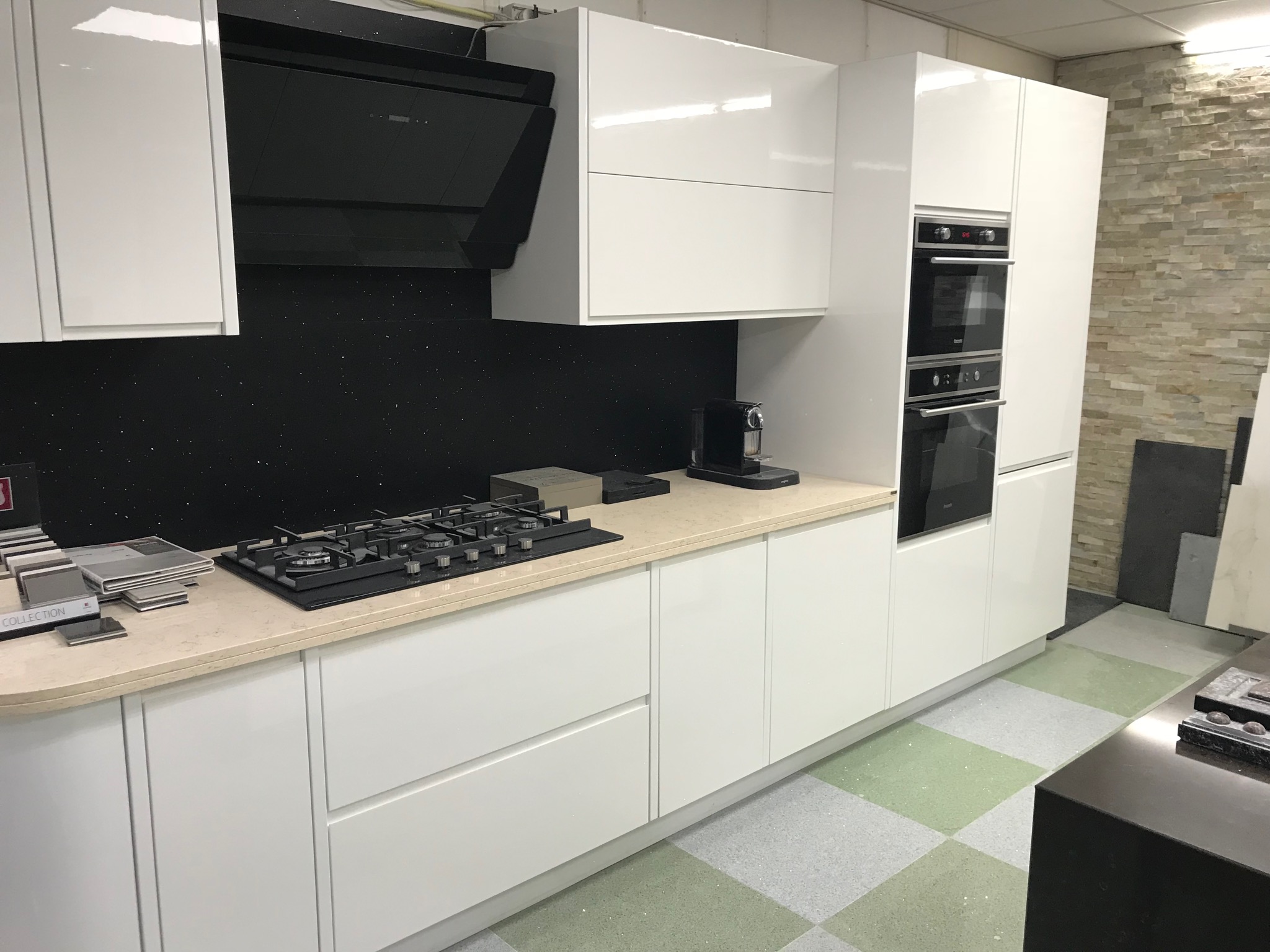 Biała, nowoczesna kuchnia z połyskiem, z czarnym panelem nad blatem imitującym nocne niebo, wyposażona w płytę gazową, piekarnik i ekspres do kawy, z blatem w kolorze beżowym.