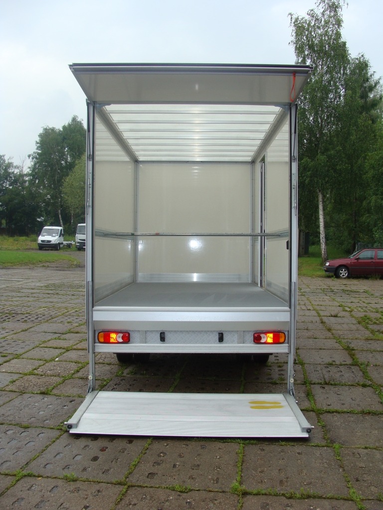 Wnętrze paki busa z otwartą rampą załadunkową, widok z tyłu, na zewnątrz teren zielony i zaparkowane samochody.