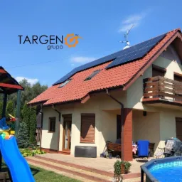 Dom jednorodzinny z panelami fotowoltaicznymi na dachu, placem zabaw i basenem w ogrodzie, logo firmy TARGEN grupa na niebie.