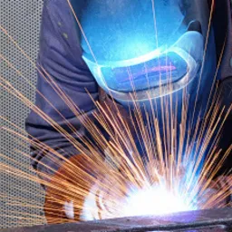 Spawacz w ochronnej masce podczas pracy, iskry rozchodzą się promieniście na tle perforowanej metalowej ściany.