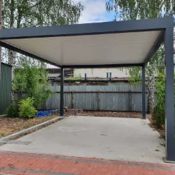 Nowoczesna, szara pergola z płaskim, białym dachem na betonowej wylewce, otoczona zielenią i ogrodzeniem z blachy falistej.