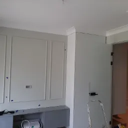 Ściana z ozdobnymi panelami i listwami w trakcie montażu, zabezpieczona niebieską taśmą malarską, z widoczną szafką pod telewizor i drabiną opartą o ścianę.