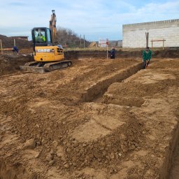 F.U.H. MAYAM - Żółta koparka CASE w wykopie pod fundamenty, obok pracownicy z łopatą i niwelatorem. W tle widać częściowo wzniesione ściany budynku.