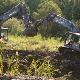 F.U.H. MAYAM - Dwie koparki Volvo pracują na nierównym terenie porośniętym trawą, w tle widoczne drzewa. Jedna koparka podnosi urobek, druga stoi na wzniesieniu.
