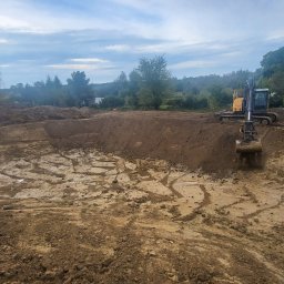 F.U.H. MAYAM - Żółto-czarna koparka podczas wykopu ziemi w Sławnie. Widoczne świeże ślady opon i łyżki koparki. Tło stanowią drzewa i pochmurne niebo.