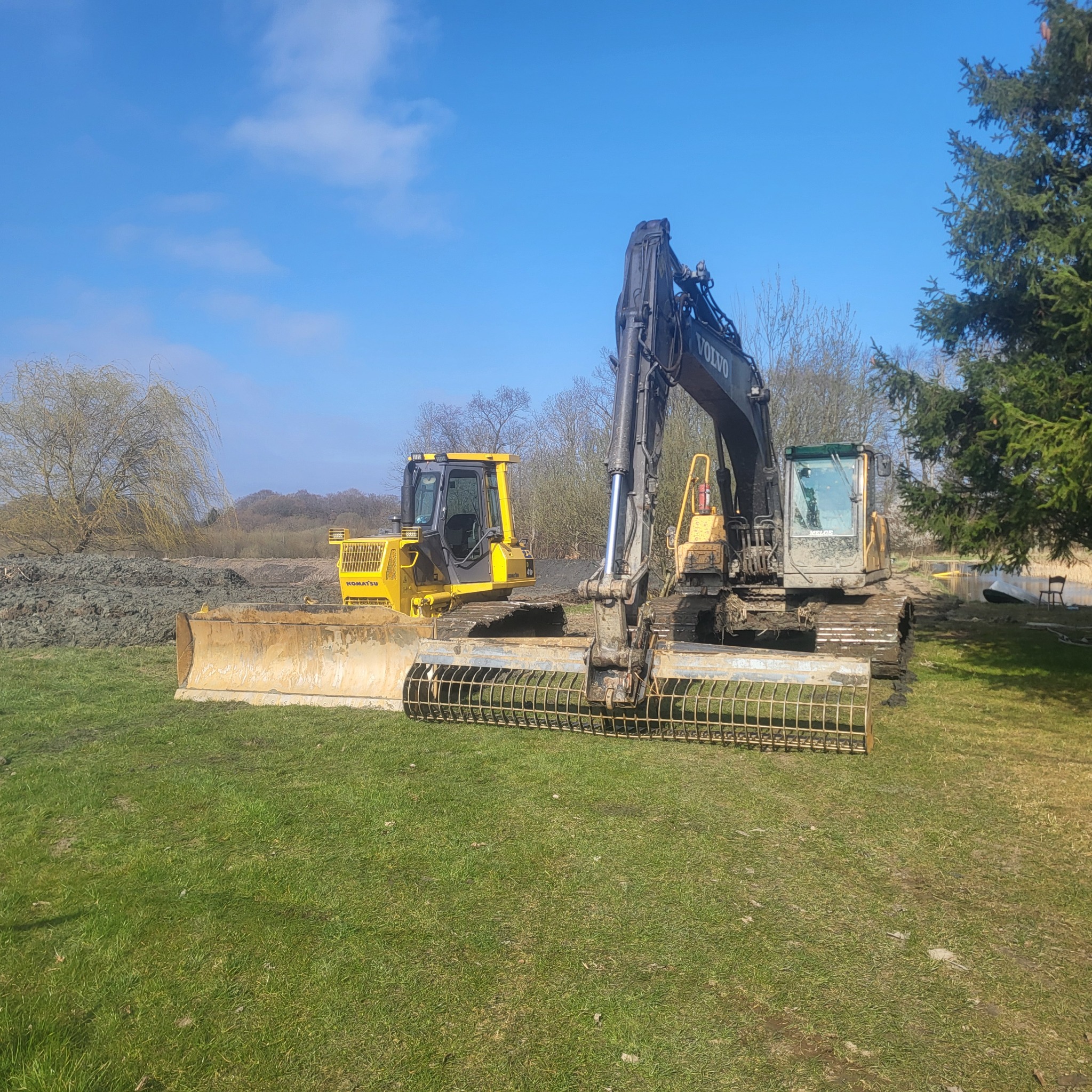 Żółty spychacz Komatsu i czarna koparka Volvo na trawie, z hałdą ziemi w tle, prawdopodobnie po niwelacji terenu.