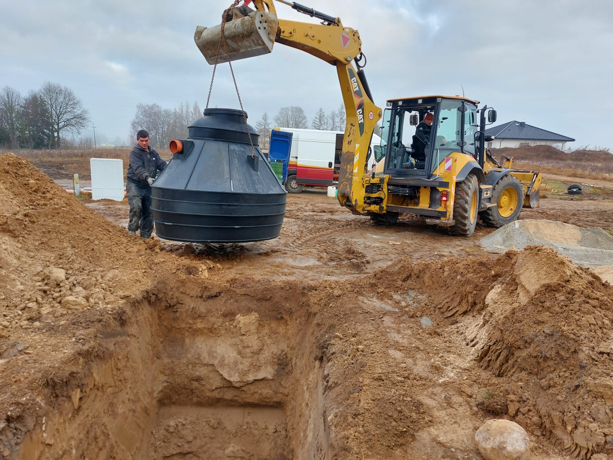 Żółta koparko-ładowarka umieszcza czarny zbiornik z tworzywa w wykopie, obok stoi pracownik. W tle budowa domu i zaparkowany bus.