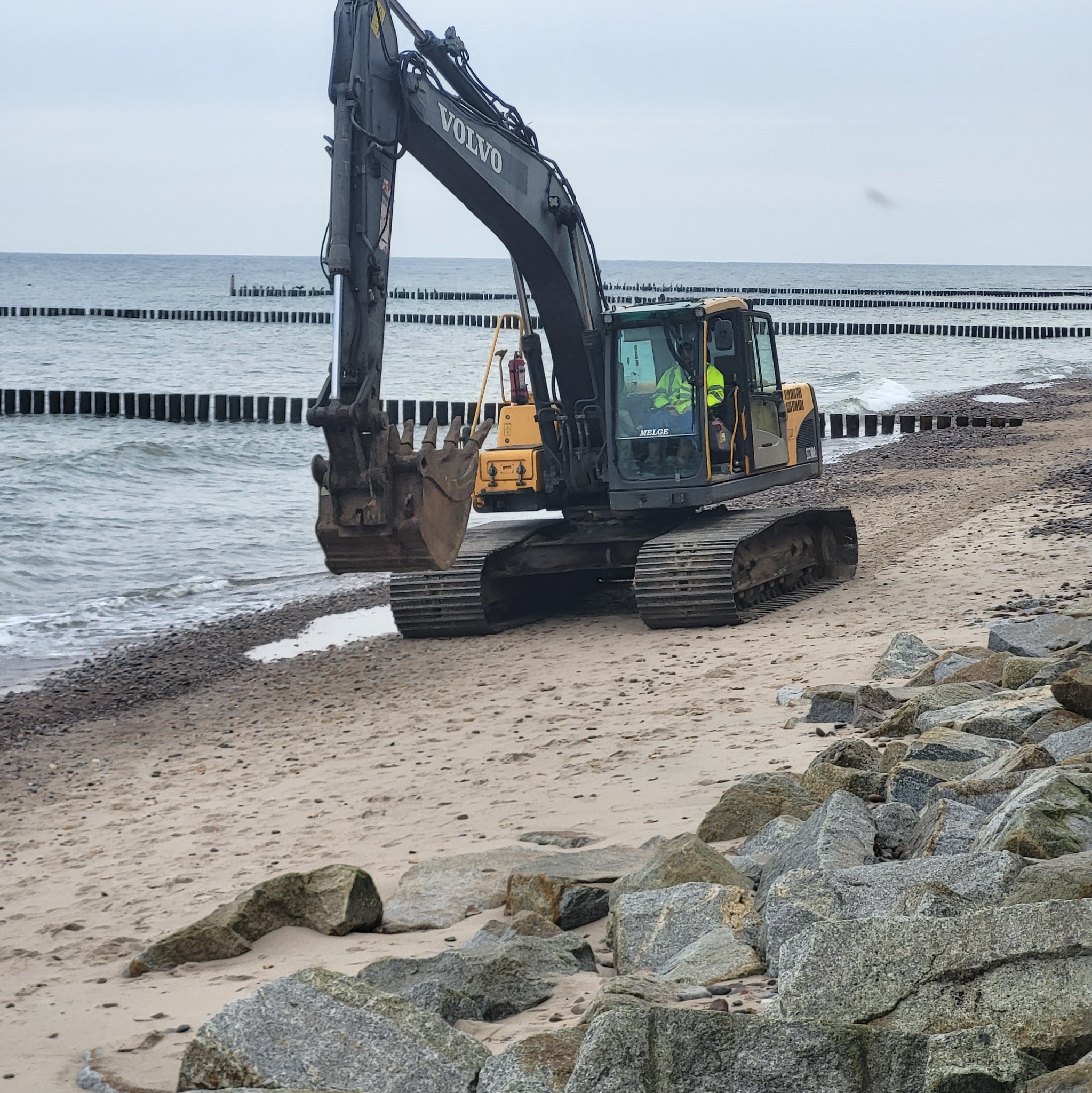 Żółto-czarna koparka Volvo na piaszczystej plaży z widokiem na morze i drewniane falochrony. Operator w kabinie. Skaliste nabrzeże na pierwszym planie.