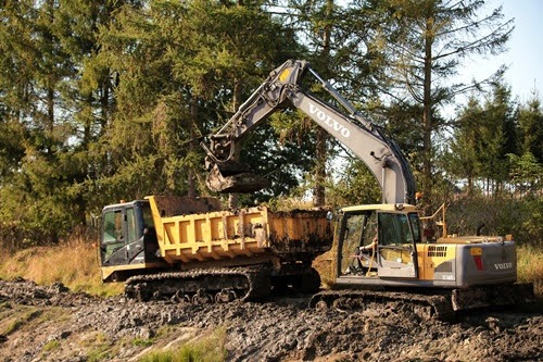 Żółto-szara koparka Volvo na gąsienicach załadowuje ziemię do wywrotki na tle drzew. Prace ziemne w terenie.