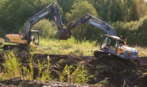 Dwie koparki Volvo pracują na nierównym terenie porośniętym trawą, w tle widoczne drzewa. Jedna koparka podnosi urobek, druga stoi na wzniesieniu.