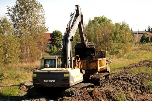 Żółta koparka Volvo nabiera ziemię do wywrotki na tle drzew i budynku. Prace ziemne w toku.