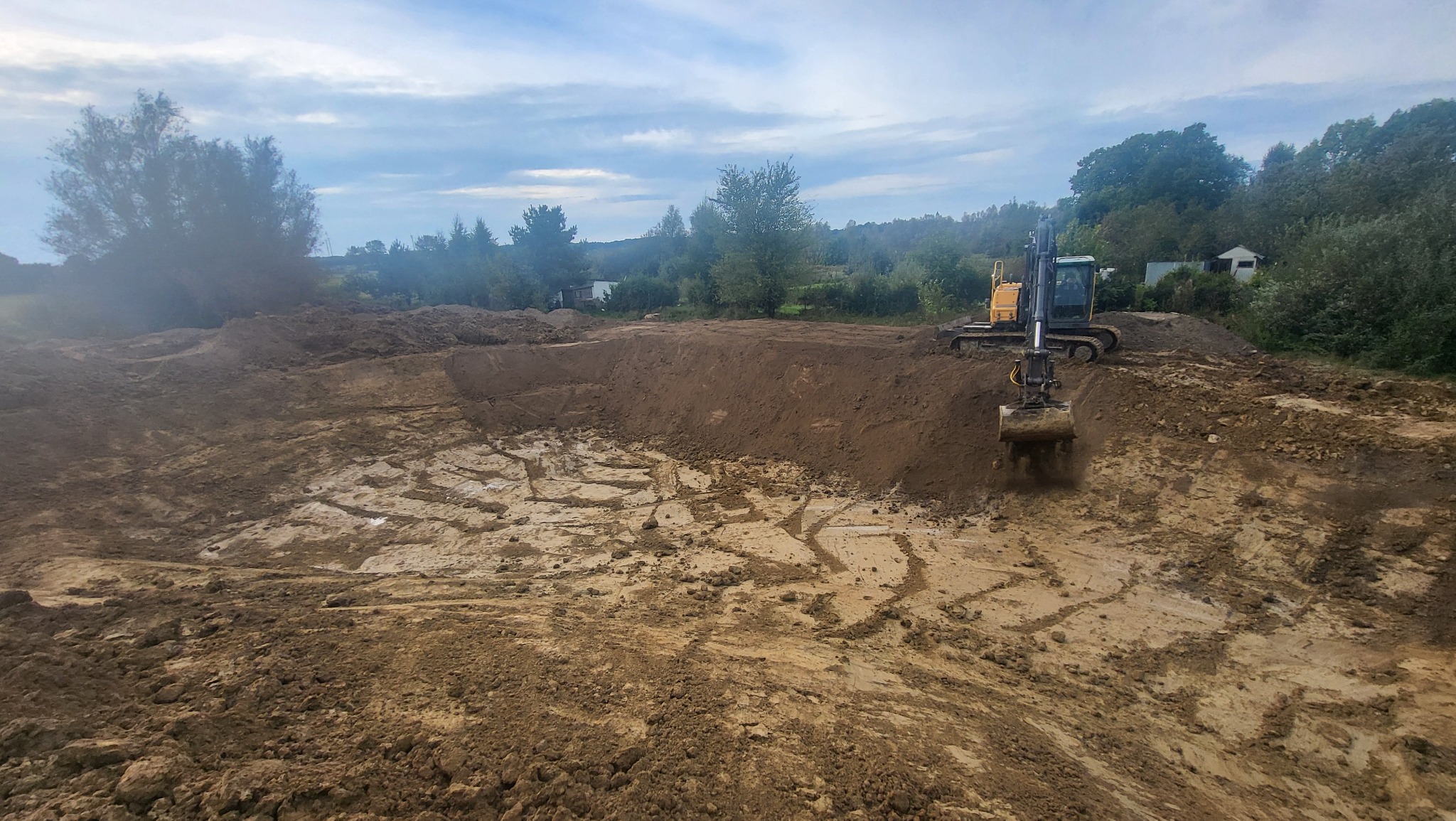 Żółto-czarna koparka podczas wykopu ziemi w Sławnie. Widoczne świeże ślady opon i łyżki koparki. Tło stanowią drzewa i pochmurne niebo.