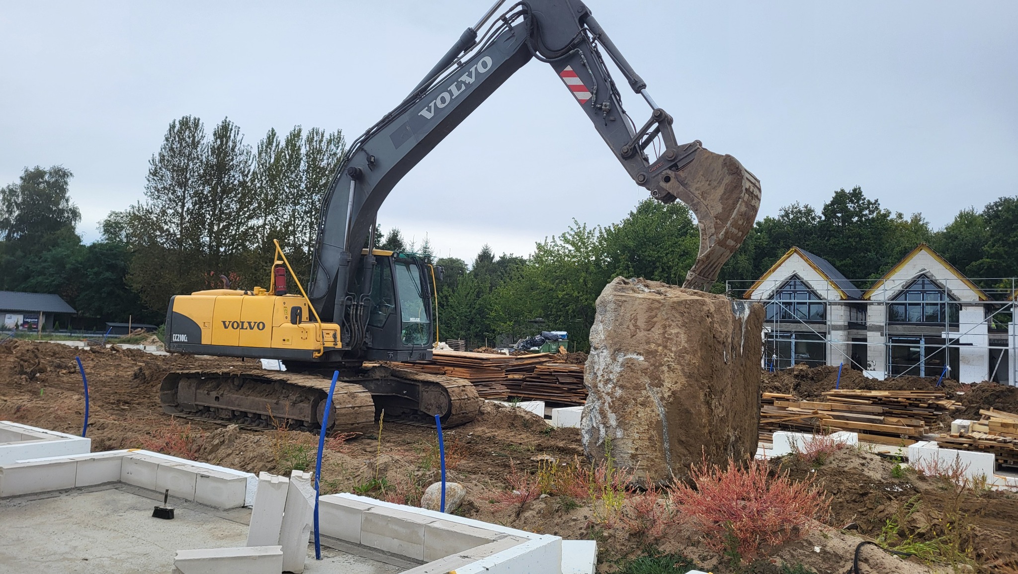 Żółta koparka Volvo podnosi duży kamień na placu budowy z fundamentami i domem w tle, na tle pochmurnego nieba.