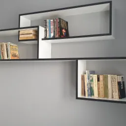 Asymetryczna, nowoczesna półka ścienna w kolorze białym z czarnym obramowaniem, wypełniona książkami o różnej wielkości i kolorystyce okładek, na tle szarej ściany i fragmentu zasłony.