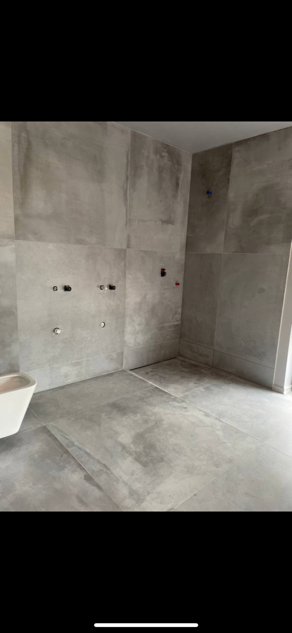 Wykończona łazienka z szarymi, wielkoformatowymi płytkami imitującymi beton, z widocznymi przyłączami wodno-kanalizacyjnymi i częściowo zamontowaną białą miską WC.