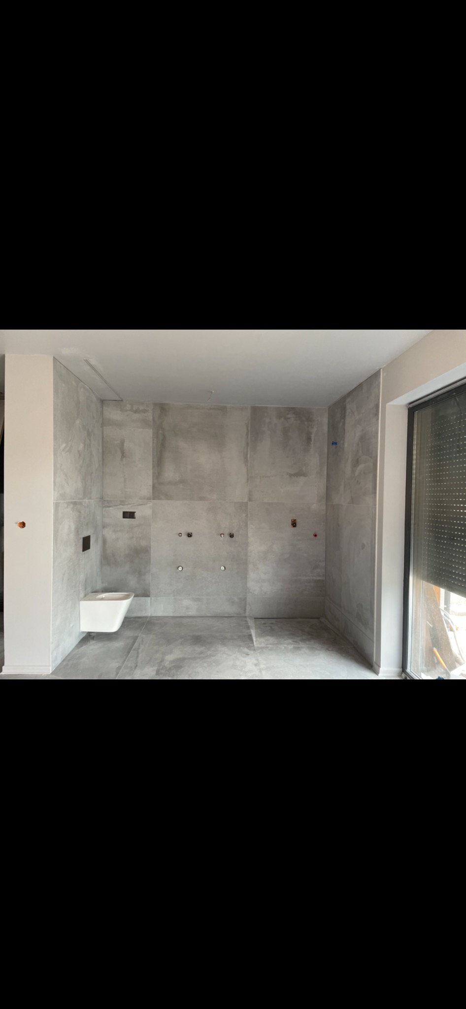 Surowa łazienka w trakcie remontu z szarymi płytkami imitującymi beton, wisząca toaleta zamontowana, widoczne instalacje wodno-kanalizacyjne przed podłączeniem armatury.