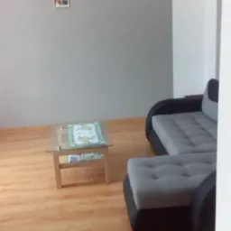 Jasne panele podłogowe w salonie z szarą ścianą, szarym narożnikiem z czarnymi bokami i szklanym stolikiem kawowym z dekoracyjną serwetą.