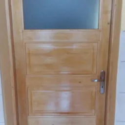 Jasne, drewniane drzwi wewnętrzne z prostokątną szybą u góry i trzema panelami poniżej, widziane z bliska, z metalową klamką.